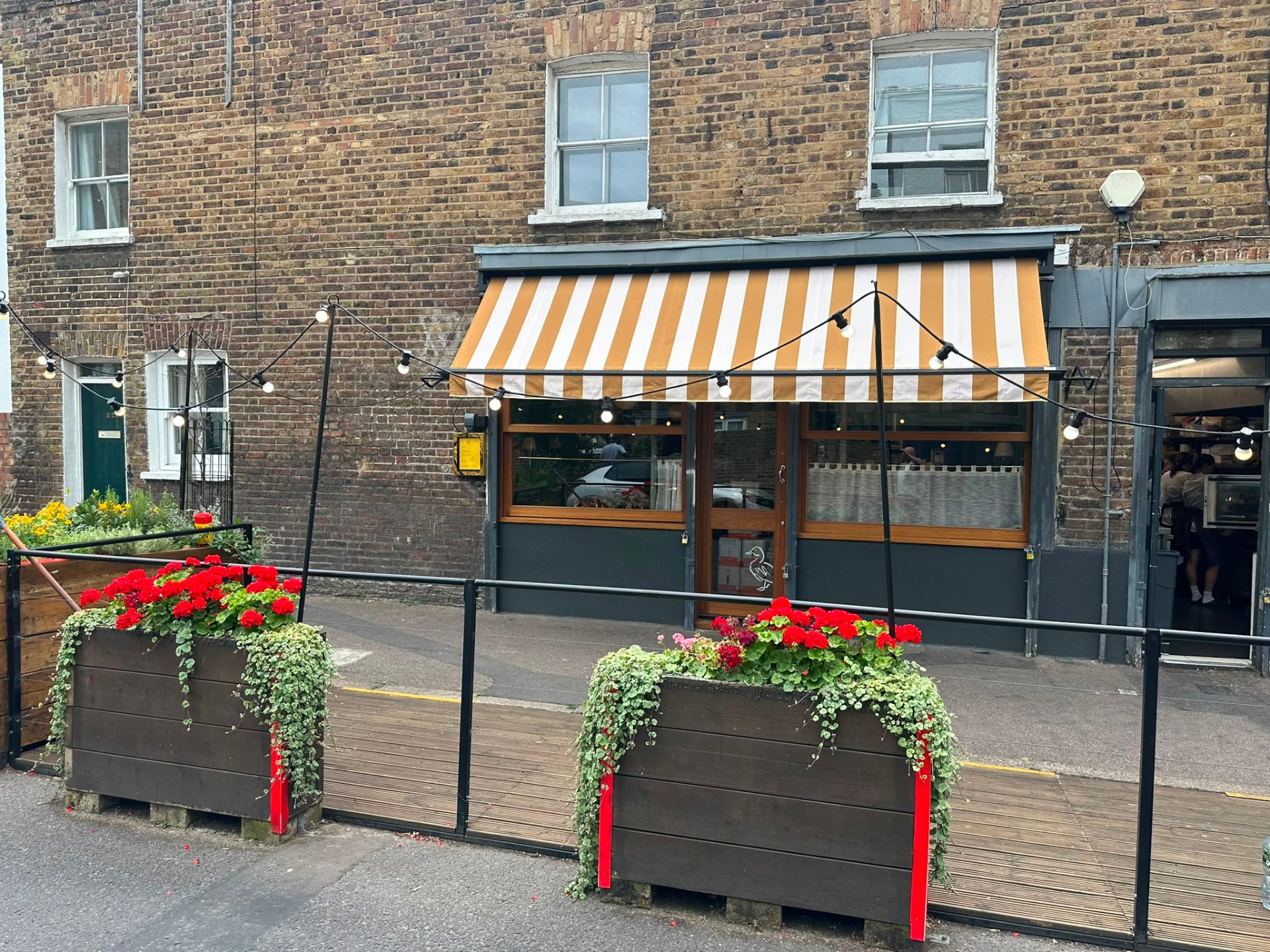 Traditional drop-arm awning in beige and cream stripe over a Hackney shopfront with planters and festoon lights